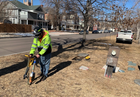 A Utility Locates team member uses a device to help locate utilities in the area. The worker is wearing a fluorescent, high-visibility work jacket and a hard hat