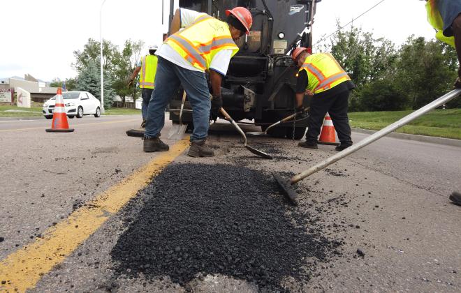 three people smooth black asphalt in a large pothole to prepare for steam rolling. A large truck that spreads the asphalt is in the background. Orange cones direct traffic away from the crew.